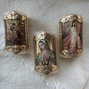 Op de afbeelding: Drie witte pilaar kaarsen met religieuze afbeeldingen. Elke kaars heeft een goudkleurige, sierlijke lijst. De afbeeldingen omvatten een engel, de Maagd Maria en Jezus. De kaarsen staan op een wit, getextureerd oppervlak.