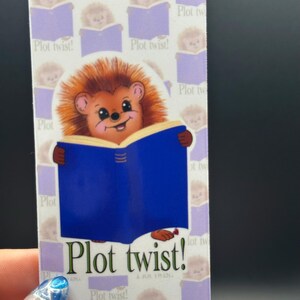 Peut inclure: Marque-page rectangulaire avec un hérisson de dessin animé lisant un livre bleu. Le hérisson a des poils bruns et un visage souriant. Le marque-page a un motif répété de hérissons et le texte "Plot twist!"