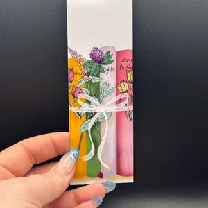 May include: A rectangular bookmark featuring three colorful book spines with floral accents and the text "Jane Austen". The spines are yellow, green, and pink, tied with a white ribbon. A small ladybug is visible.