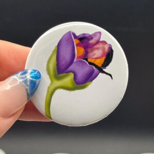 Può includere: Un bottone bianco con un design floreale. L'immagine presenta un fiore viola e arancione con uno stelo verde. Il bottone è tenuto in mano con un design per unghie blu e argento. Lo sfondo è nero.