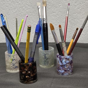 May include: Three cylindrical resin pen holders filled with various art supplies. One holder contains dark brown stone chips, another features clear translucent chips, and the third is filled with purple and blue stone fragments.