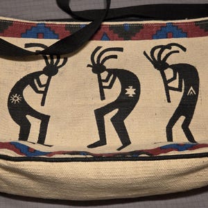 May include: A rectangular woven shoulder bag in cream, featuring three black Kokopelli figures playing flutes. The bag has a black strap and a border pattern in red, blue, and black. The figures include small white symbols on their bodies.