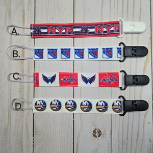May include: Four different pacifier clips with various hockey team logos. The clips are white, black, and red with blue accents. The logos include the New York Rangers, Washington Capitals, and New York Islanders.