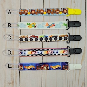 May include: Five different types of colorful fabric ribbon with a clip attached to the end. The ribbons feature various designs including cowboys, airplanes, monster trucks, NASCAR, and Hot Wheels.