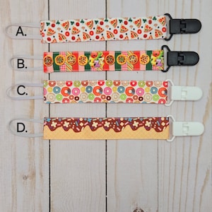 May include: Four different pacifier clips with various food-themed patterns. The clips are white, black, and white. The patterns include pizza, donuts, and a chocolate-covered donut with sprinkles.