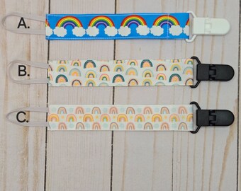 May include: Three pacifier clips with different rainbow patterns. The top clip has a blue background with white clouds and rainbow arches. The middle clip has a white background with a rainbow arch pattern. The bottom clip has a white background with a rainbow arch pattern and small dots. All clips have a white elastic loop and a black or white plastic clip.