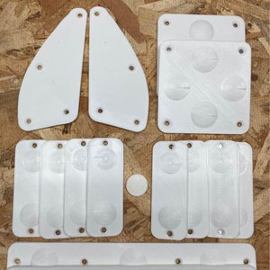 May include: A collection of white plastic components, including two curved pieces, several rectangular plates with circular designs, and a long, rounded piece with evenly spaced holes. The pieces are arranged on a wooden surface.