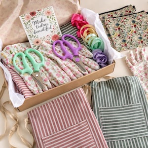 May include: A gift box containing floral print gardening gloves, a pair of mint green scissors, a pair of purple scissors, and a card with the text Make today beautiful. Also included are two striped aprons, two floral pouches, and ribbons.