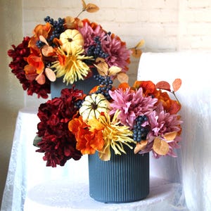 Könnte beinhalten: Zwei Blumengestecke in tealblauen, geriffelten Vasen. Die Arrangements zeigen eine Mischung aus Herbstfarben, darunter bordeauxrote, orangefarbene, gelbe und rosa Blüten, mit kleinen weißen Kürbissen und dunkelblauen Beeren. Die Arrangements stehen auf einer weißen Oberfläche.