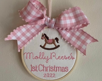 May include: Embroidered Christmas ornament with a pink and white gingham bow. The ornament features a rocking horse design and the text "Molly Reese's 1st Christmas 2022" in pink thread, set in a wooden hoop.