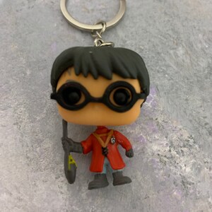 May include: A black and red Harry Potter character keychain with a black wand and round glasses. The character is wearing a red robe with a yellow symbol on the front.