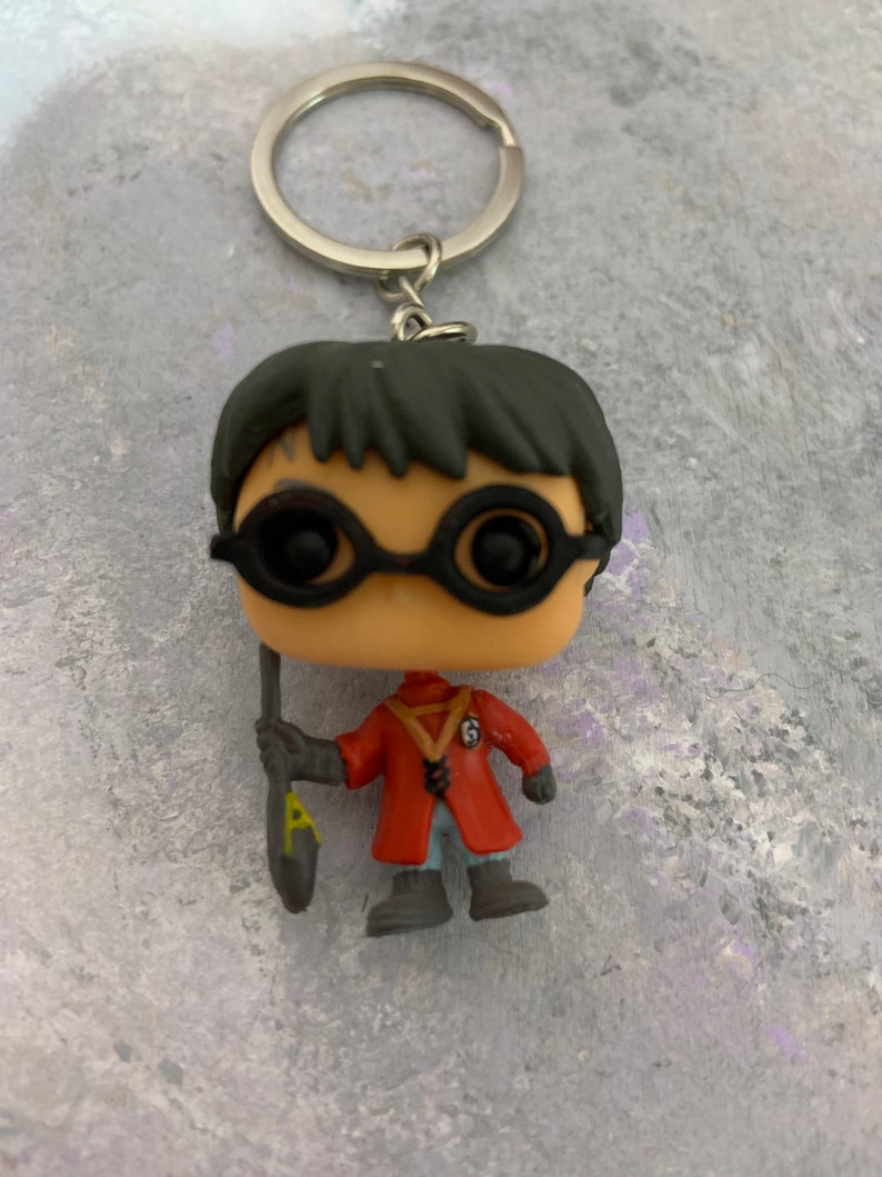 May include: A black and red Harry Potter character keychain with a black wand and round glasses. The character is wearing a red robe with a yellow symbol on the front.
