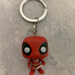 May include: A red and black Deadpool character keychain. The keychain is a small, stylized figure of Deadpool with a silver knife in his right hand. The keychain is attached to a silver metal ring.