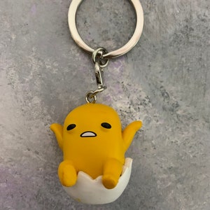 May include: A yellow cartoon egg character keychain with a white eggshell. The character has black eyes and a surprised expression. The keychain is attached to a silver metal ring.