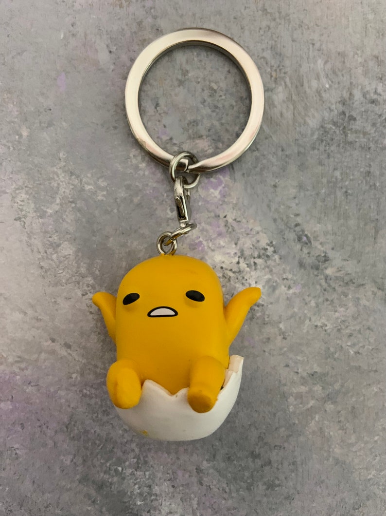 May include: A yellow cartoon egg character keychain with a white eggshell. The character has black eyes and a surprised expression. The keychain is attached to a silver metal ring.