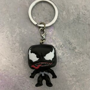 May include: A black and white Venom keychain with a red tongue. The keychain is a small, stylized figure of the Marvel Comics character Venom. The figure is attached to a silver key ring.