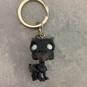 May include: A black and white keychain featuring a small figurine of a griffin. The griffin has a black body and wings, a brown beak, and white eyes. The keychain is attached to a silver metal ring.