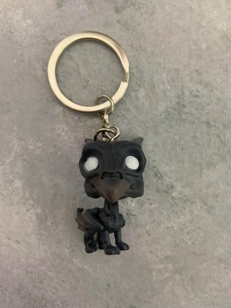 May include: A black and white keychain featuring a small figurine of a griffin. The griffin has a black body and wings, a brown beak, and white eyes. The keychain is attached to a silver metal ring.
