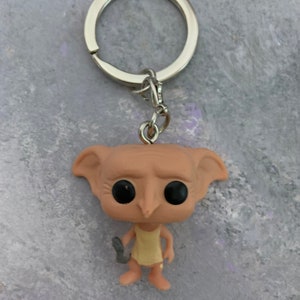 May include: A Dobby the house-elf keychain with a silver key ring. Dobby is wearing a yellow dress and holding a sock in his left hand. He has large black eyes and a pointed nose.