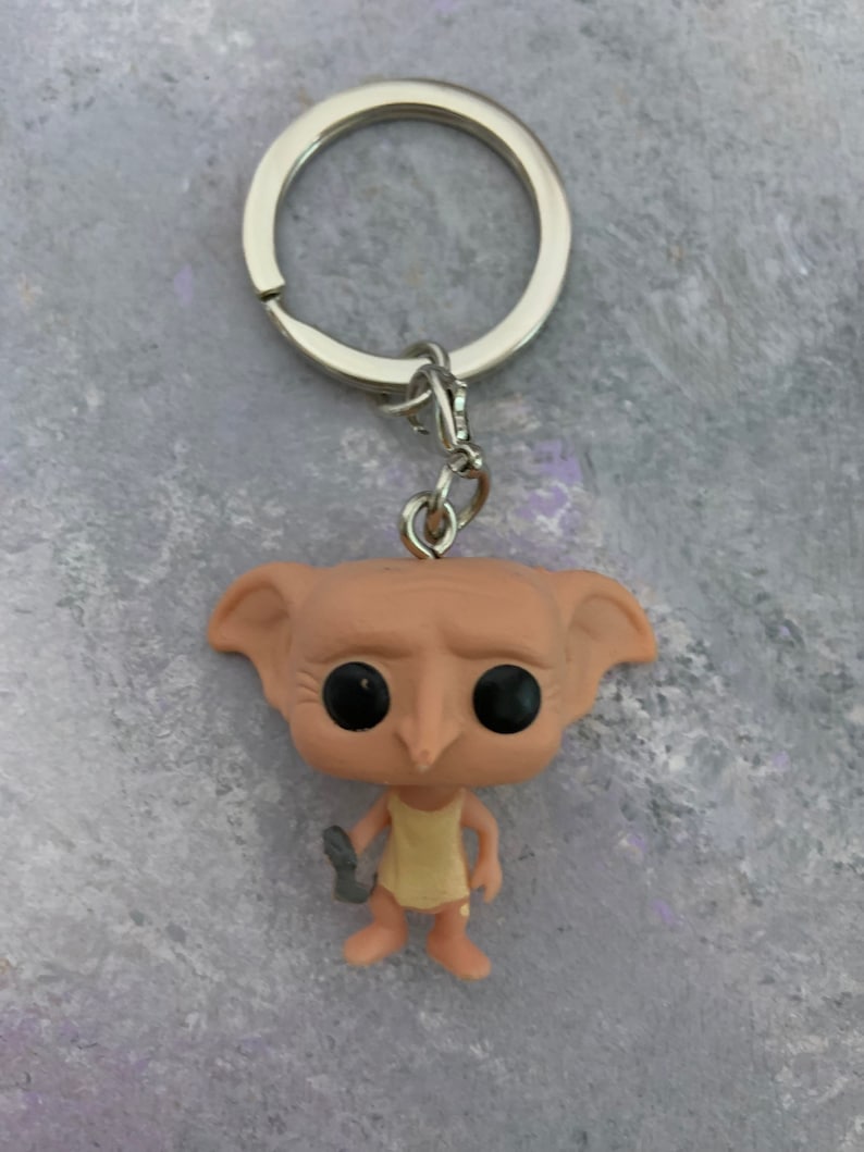 May include: A Dobby the house-elf keychain with a silver key ring. Dobby is wearing a yellow dress and holding a sock in his left hand. He has large black eyes and a pointed nose.