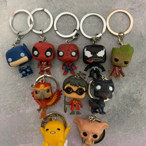 May include: A collection of ten keychains featuring various characters. The keychains are made of plastic and feature characters such as Batman, Deadpool, Spiderman, Venom, Groot, a phoenix, Harry Potter, a dragon, a yellow character with a white egg, and a pink character with large ears.