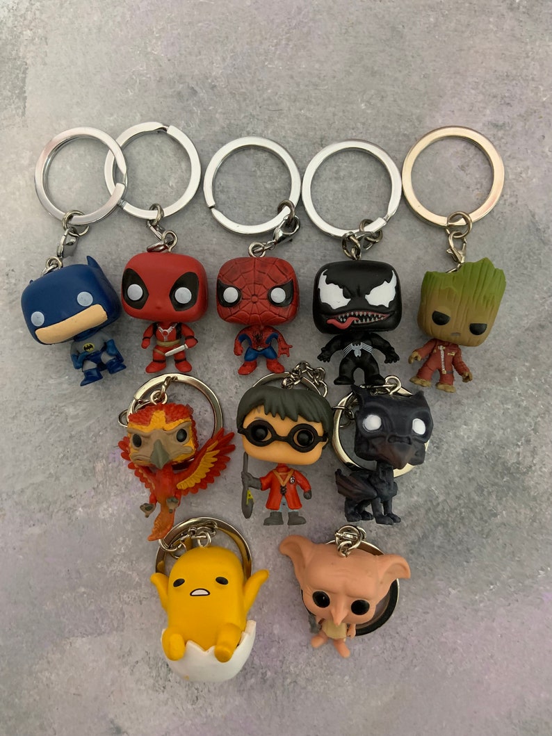May include: A collection of ten keychains featuring various characters. The keychains are made of plastic and feature characters such as Batman, Deadpool, Spiderman, Venom, Groot, a phoenix, Harry Potter, a dragon, a yellow character with a white egg, and a pink character with large ears.