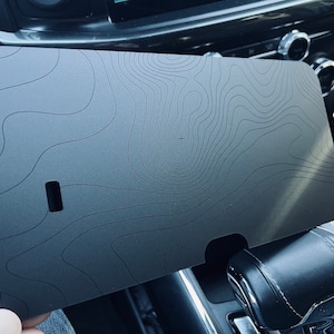 May include: A gray, rectangular, topographic map-style phone holder for a car. The holder has a small rectangular cutout in the center.