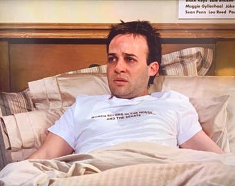May include: A man in a white t-shirt with the text "WOMEN BELONG IN THE HOUSE... AND THE SENATE" printed on it, lying in bed with a white blanket and striped pillows.