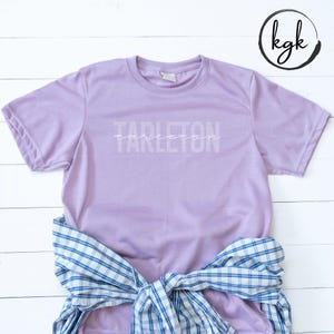 Könnte beinhalten: Ein lavendelfarbenes T-Shirt mit dem Wort "Tarleton" in weiß auf der Vorderseite. Das Shirt liegt flach auf einer weißen Holzoberfläche, um die ein blau-weiß kariertes Hemd gebunden ist.