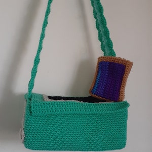 May include: A green crocheted bag with a white and black trim. The bag has a long green crocheted strap and a small purple and brown crocheted pouch attached to the front.