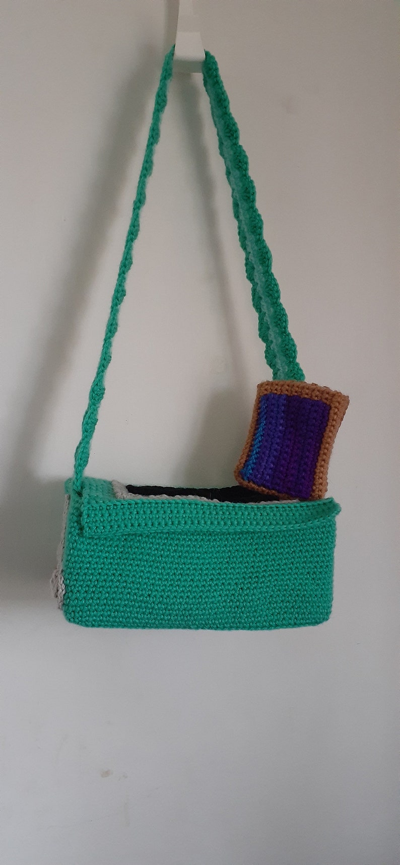 May include: A green crocheted bag with a white and black trim. The bag has a long green crocheted strap and a small purple and brown crocheted pouch attached to the front.