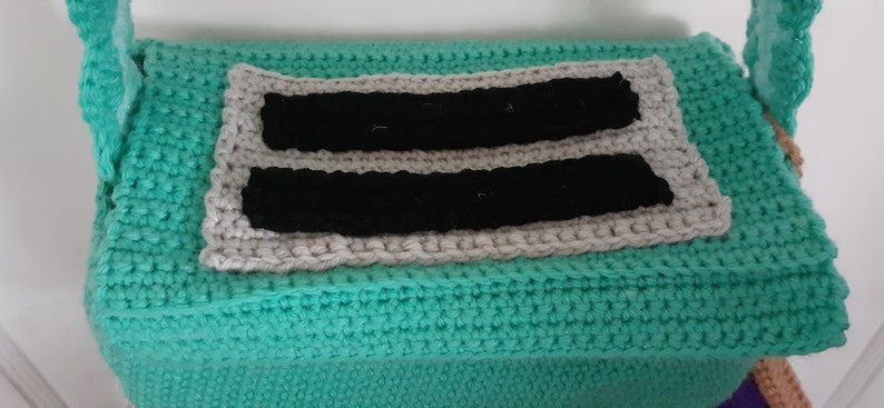May include: A teal crocheted bag with a gray and black crocheted panel on the front. The panel has two black stripes.