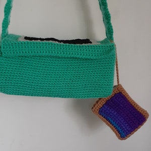 May include: A green crocheted bag with a black and white design on the front. A small purple and brown crocheted pouch is attached to the bag.