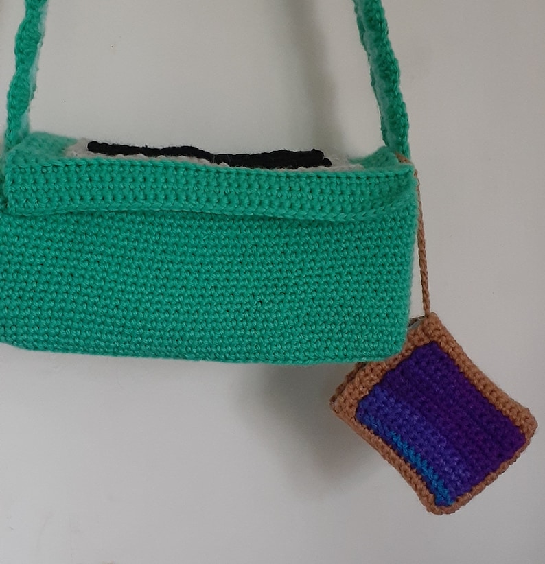 May include: A green crocheted bag with a black and white design on the front. A small purple and brown crocheted pouch is attached to the bag.
