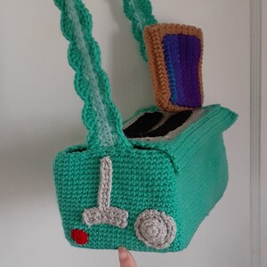 May include: A green crocheted toaster bag with a brown crocheted bread slice inside. The bag has a gray crocheted handle and a red crocheted button.