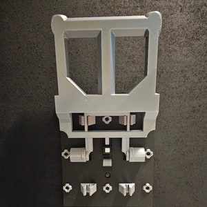 May include: A gray 3D-printed object with a rectangular base and various components, including a frame, cylinders, and small, star-shaped pieces. The object is set against a dark background.