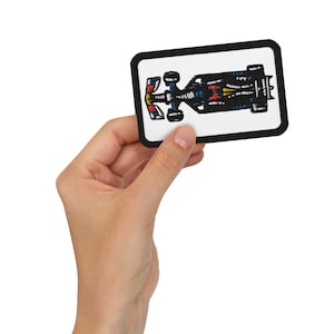 Puede incluir: Un parche bordado en blanco y negro de un coche de carreras con detalles rojos, azules y amarillos. El parche está sobre un fondo blanco con un borde negro.