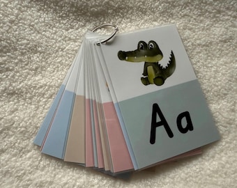 Puede incluir: Un conjunto de tarjetas didácticas del alfabeto sujetas por un anillo metálico. Cada tarjeta presenta una letra y una ilustración correspondiente, como la letra "A" con un cocodrilo de dibujos animados. Las tarjetas tienen acentos de color pastel.