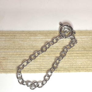 May include: A silver-colored chain bracelet with an oval link design and a toggle clasp. The bracelet is displayed on a white surface with a beige textured background.