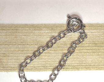 May include: A silver-colored chain bracelet with an oval link design and a toggle clasp. The bracelet is displayed on a white surface with a beige textured background.