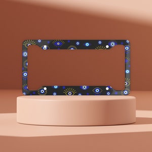 May include: Black license plate frame with a pattern of blue and gold evil eyes. The frame is on a light pink surface.