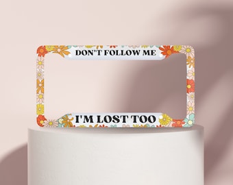 May include: White license plate frame with a floral design in shades of orange, yellow, pink, and green. The text "Don't Follow Me I'm Lost Too" is printed on the frame.