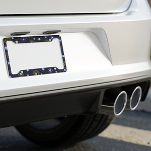 May include: A black license plate frame with a white border and a pattern of blue and yellow evil eyes. The frame is attached to the back of a white car.