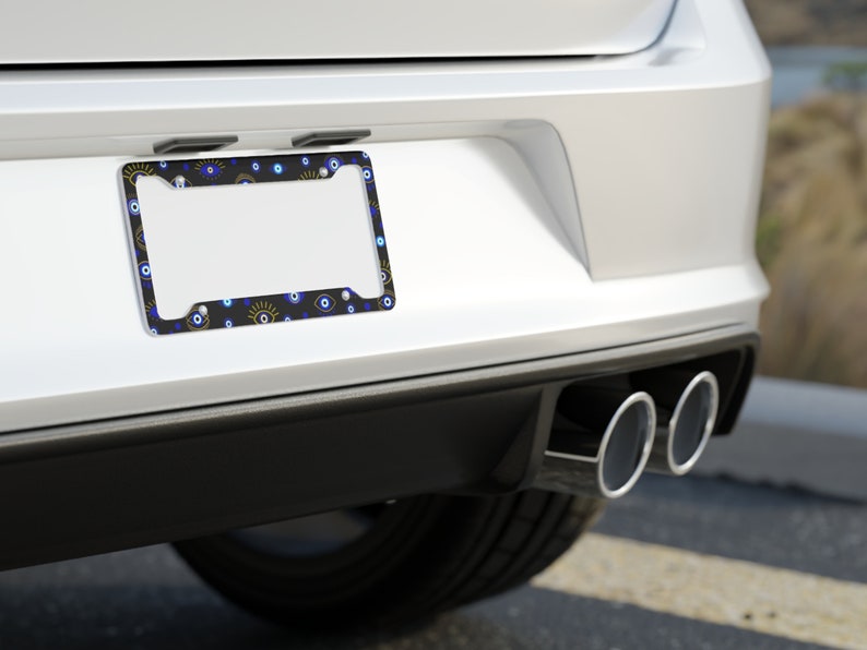 May include: A black license plate frame with a white border and a pattern of blue and yellow evil eyes. The frame is attached to the back of a white car.