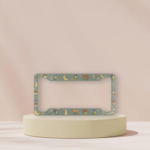 May include: A license plate frame featuring a pattern of mushrooms, moons, and stars on a teal background. The rectangular frame has rounded corners and is designed to fit a standard UK license plate.