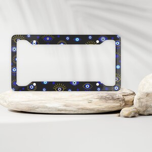 May include: A black license plate frame with a pattern of blue and gold evil eyes. The frame is on a wooden surface.