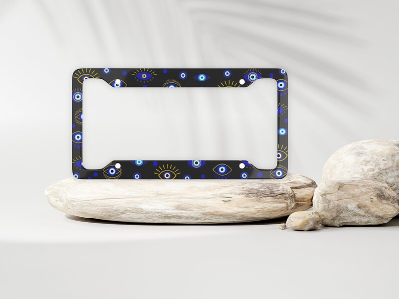 May include: A black license plate frame with a pattern of blue and gold evil eyes. The frame is on a wooden surface.