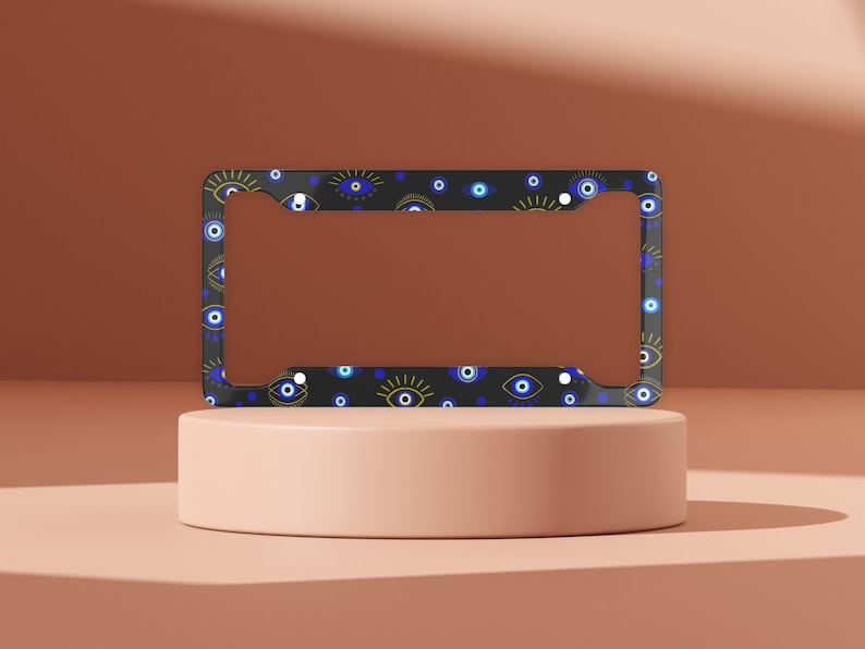 May include: Black license plate frame with a pattern of blue and gold evil eyes. The frame is on a light pink surface.