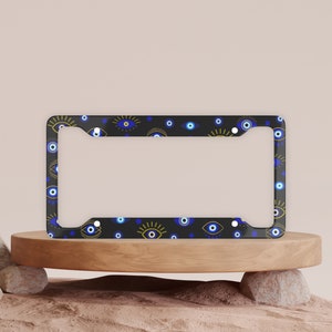 May include: A black license plate frame with a pattern of blue and gold evil eyes. The frame is on a wooden platform.