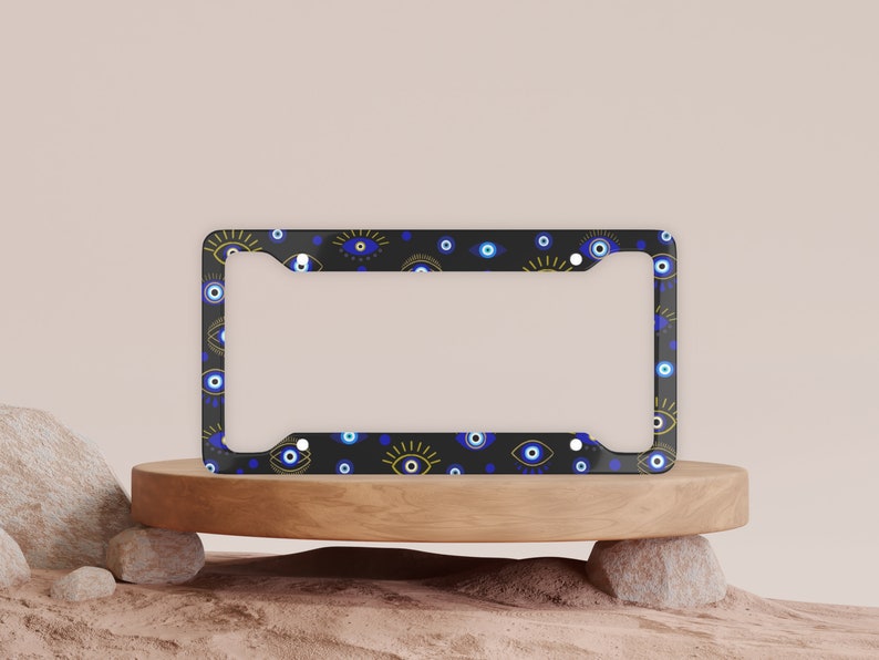 May include: A black license plate frame with a pattern of blue and gold evil eyes. The frame is on a wooden platform.